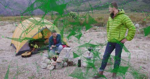 Crouching woman tending campfire while partner observing rugged desert campsite with tent