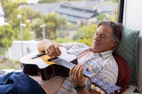 Senior man relaxing and playing acoustic guitar on sunlit window seat at home