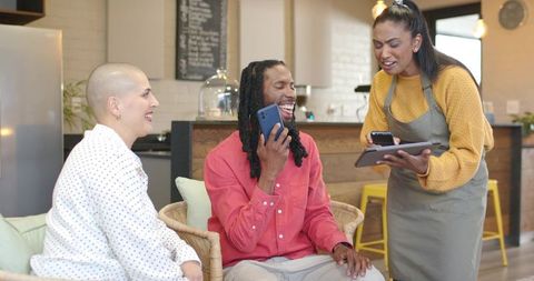 Laughing diverse friends ordering and chatting in cozy cafe while barista uses tablet