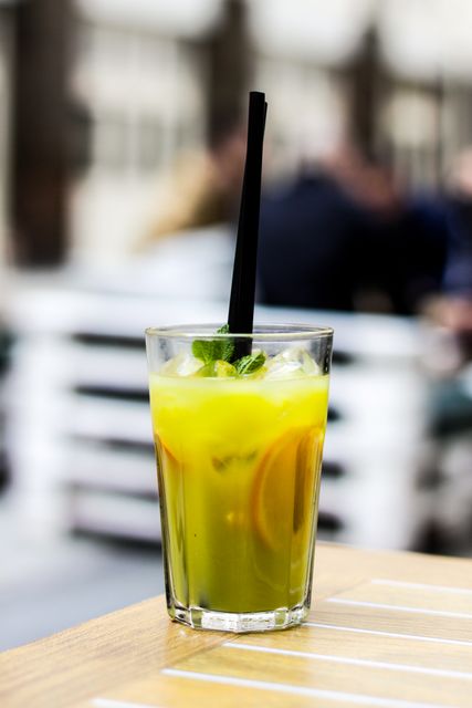 Lemon drink with mint leaves and ice in a clear glass with a black straw, placed on a wooden table with a blurred background. Ideal for illustrating refreshing summer beverages, outdoor dining experiences, or health-conscious drink options. Suitable for use in advertising, cafe menu design, and lifestyle blogs.