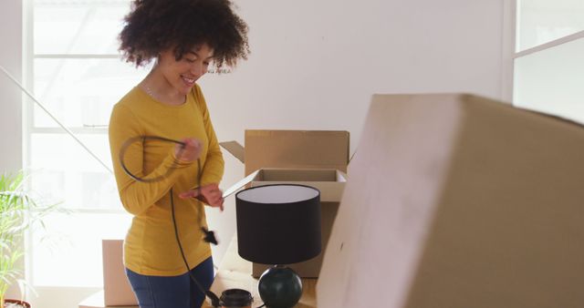 Young woman smiling while unpacking boxes in a new apartment. She is holding a power cord and a lamp is on the table nearby. Useful for portraying themes of relocation, new beginnings, home organization, and setting up a new living space.