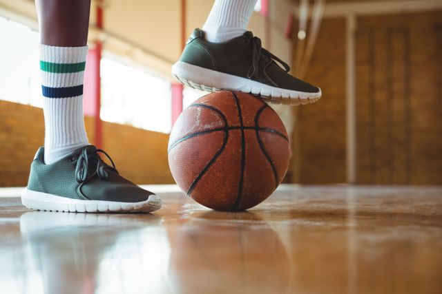 This image shows a teenager's legs and feet, with one foot stepping on a basketball in a gym. The teenager is wearing athletic sneakers and striped socks. This image can be used for sports-related content, fitness blogs, basketball training programs, or youth athletic promotions.