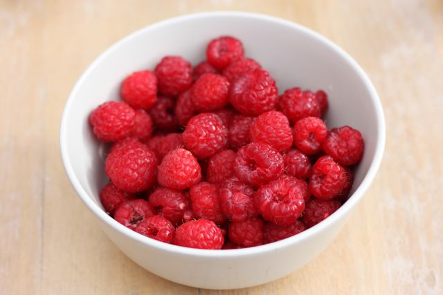 High-quality image of fresh raspberries placed in a white bowl on a wooden table. Perfect for use in health and wellness blogs, cooking websites, nutrition articles, food advertisements, product packaging, natural and organic product promotions, and diet-related content.