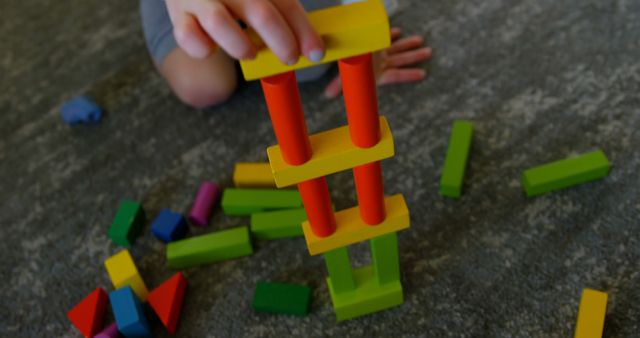 Child's hands are stacking vibrant wooden blocks on gray carpet floor. This image implies development of fine motor skills, creativity, and educational play activities. It could be used for illustrating children's classrooms, promoting educational toys, or in content about child development and early learning.