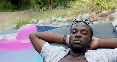 Relaxing young man lounging poolside on sunbed with sunglasses and pink float in backyard