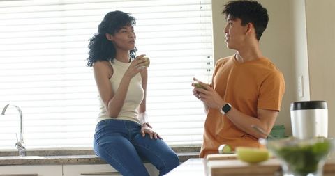 Couple enjoying green smoothie in bright kitchen, sitting on counter and chatting together