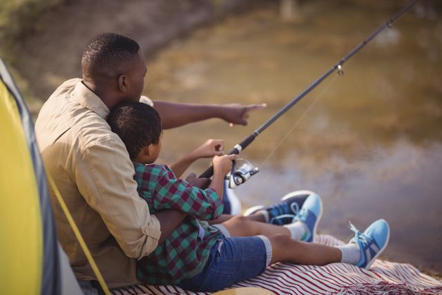 A father is showing his son how to fish by a lake in a park. They are sitting together on a blanket, the boy holding the fishing rod while the father points toward the water. Ideal for concepts of family bonding, quality time, outdoor leisure activities, and parent-child relationships. Perfect for use in advertisements, articles on family life, brochures for outdoor activities, and parenting blogs.