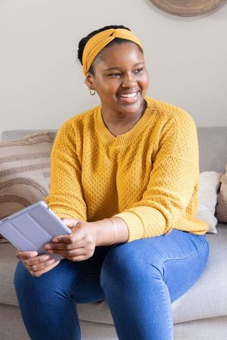 Smiling african american woman wearing mustard knit sweater and headband holding tablet on couch