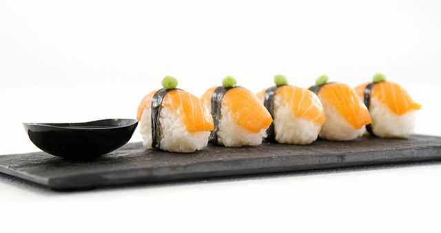 A row of sushi featuring slices of fresh salmon on top of rice, garnished with wasabi, presented on a black slate with a small bowl for soy sauce, with copy space. Sushi is a popular Japanese cuisine known for its delicate flavors and artistic presentation.