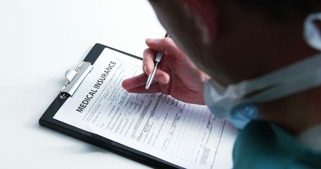 A healthcare professional, possibly a doctor or nurse, is filling out a medical insurance form on a clipboard. This image can be used in articles or websites related to health insurance, healthcare policies, medical billing, insurance claims processing, and patient care documentation.