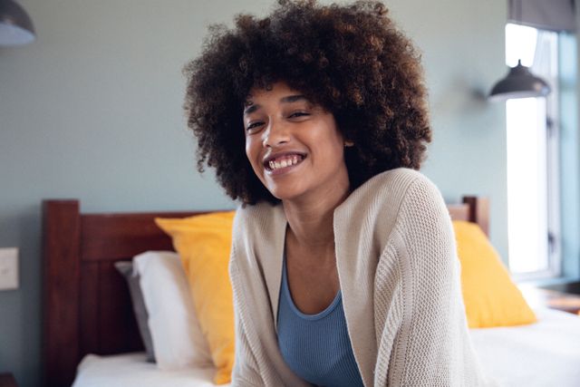 Portrait of smiling biracial young woman with afro hair sitting on bed against wall, copy space - Download Free Stock Photos Pikwizard.com