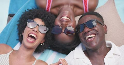 Three african american friends laughing by pool on striped towel wearing sunglasses, summer