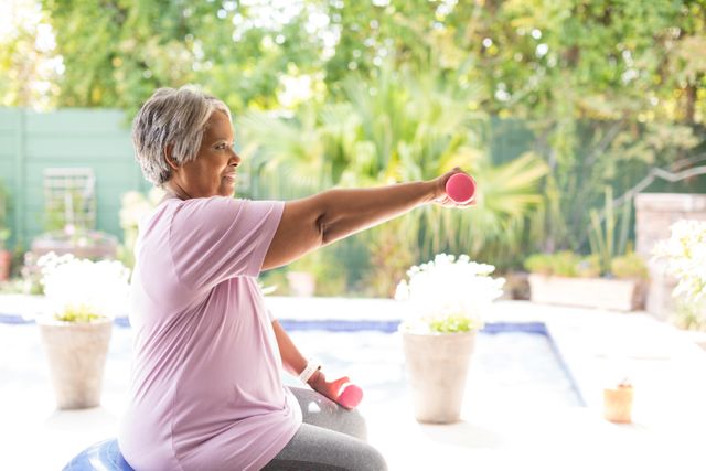 Elderly woman engaging in strength training exercises using dumbbells while sitting on a fitness ball in a garden. Ideal for promoting senior fitness, active aging, and outdoor workout concepts. Suitable for health and wellness articles, fitness blogs, and advertisements for senior exercise programs.