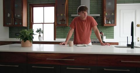 Young man leaning on modern kitchen island wearing headphones with notebook and coffee mug