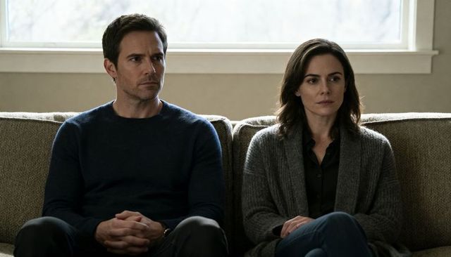 Somber couple sitting side-by-side on sofa, hands clasped, minimalist natural-light room