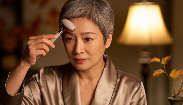 Mature woman applying rose quartz facial roller wearing satin robe in evening skincare ritual