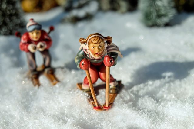 Two miniature skiers navigating through a snow-covered landscape. One skier is in the foreground, bent forward on skis with a focused expression, while the other is in the background. Great for use in winter sports promotions, holiday decoration themes, or toy collections.