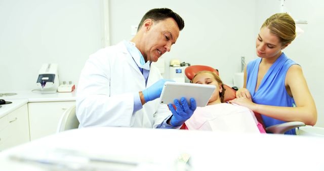 Male dentist explaining treatment plan to woman and her child using a tablet in modern dental clinic. Mother supporting daughter during dental visit. Ideal for articles on dental health, family healthcare, medical technology, dentist-patient interaction, and pediatric dentistry.