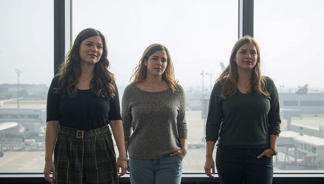 Three women standing at airport window overlooking jet bridges and terminal interiors