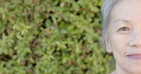 Senior asian woman smiling in garden with soft gaze and leafy green background, half portrait