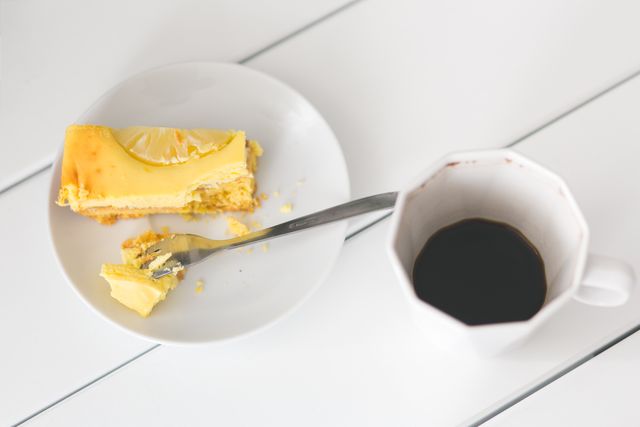 Lemon cheesecake slice on small white plate accompanied by a cup of black coffee. Cake partially eaten with fork resting on the plate. White table presents clean and minimalistic look. Ideal for foodie blogs, cafés, restaurant menus, or lifestyle articles focusing on desserts or coffee breaks.