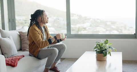 Man holding mug on sofa, looking out window in bright minimalist living room