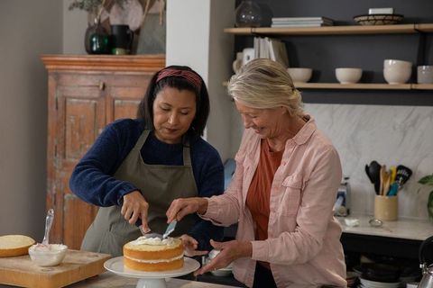 Mature friends frosting rustic vanilla layer cake in cozy home kitchen