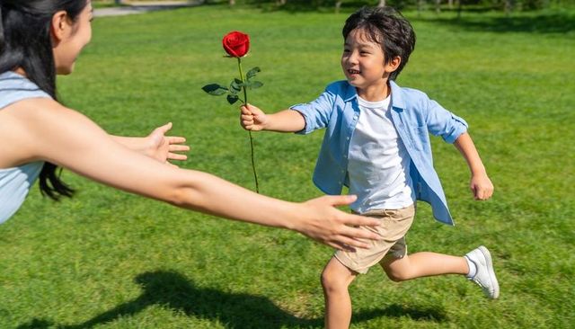 Young boy running across sunlit park presenting single red rose to woman reaching for hug