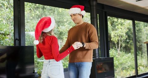 Couple dancing in cozy living room wearing santa hats by fireplace and garden windows
