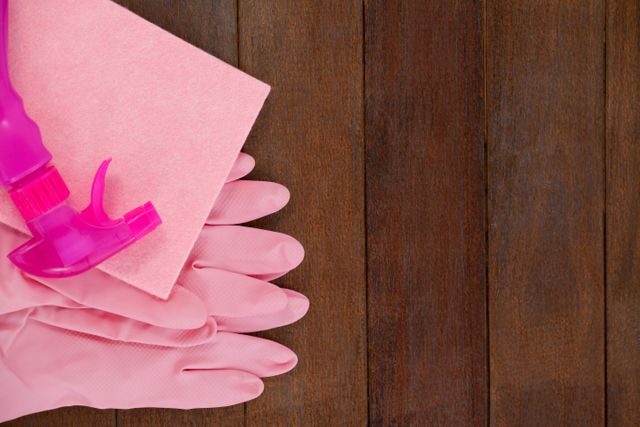 Cleaning supplies including a pink spray bottle, sponge, and gloves are arranged on a wooden floor. This can be used for content related to household cleaning, sanitation tips, and hygiene practices.
