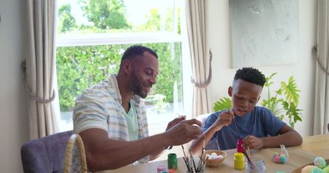 African american father and son painting easter eggs at sunlit wooden table