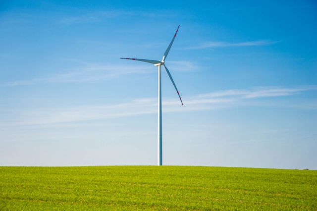 Wind turbine standing tall on a bright green field, set against a clear blue sky. Ideal for illustrating renewable energy concepts, sustainability, and eco-friendly practices. Perfect for use in environmental campaigns, energy sector publications, and green technology marketing materials.