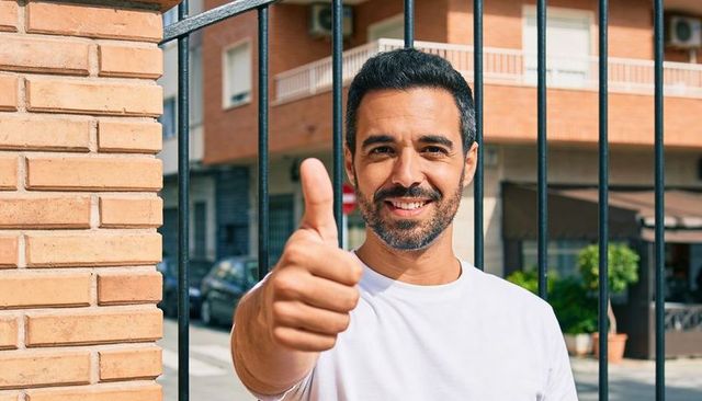 Smiling man giving thumbs up on sunny urban street near brick wall and balcony