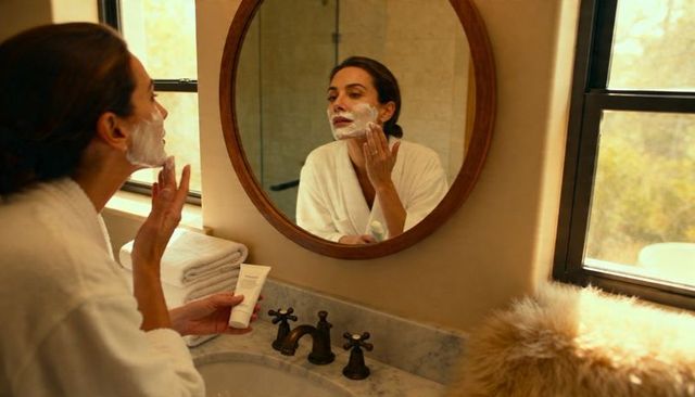 Applying facial cleanser at marble vanity wearing white robe with mirror reflection