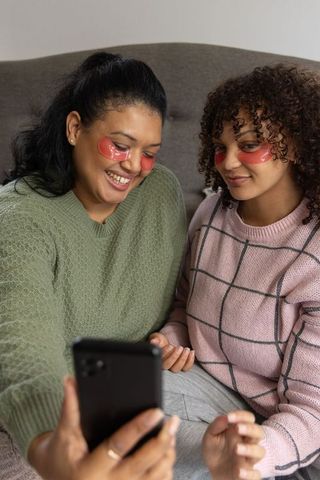 African american women sharing skincare moment with red under-eye patches and phone selfie