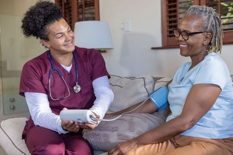Home caregiver checking blood pressure for smiling african american senior woman on couch