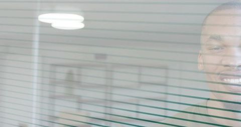 Smiling man appearing through glass with teal blinds in minimalist office interior