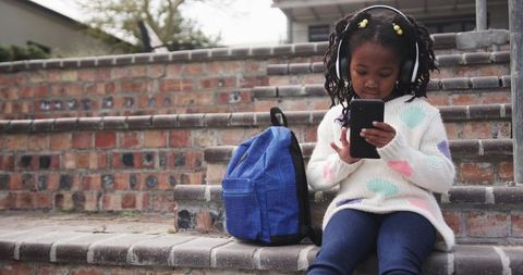This depicts a young African American girl seated on outdoor brick steps using a smartphone while wearing headphones and has a blue backpack beside her. Captures themes of modern childhood, technology use, and casual outdoor moments. Ideal for illustrating technology influence on young people, children enjoying screen time, or urban childhood scenarios.