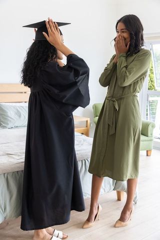 Diverse sisters celebrating graduation at home, adjusting cap and sharing joyful surprise