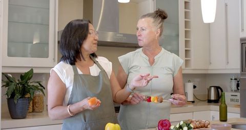 Two senior women preparing skewers together in modern kitchen
