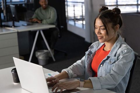 Asian woman in denim jacket typing on laptop in open-plan office with diverse coworkers