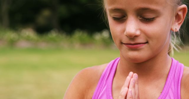 A young Caucasian girl in a pink top is practicing meditation or prayer outdoors, with copy space. Her eyes are closed, and hands pressed together in a gesture of peace or spirituality.