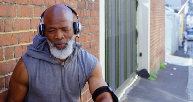 Senior man with gray beard wearing headphones while exercising outdoors. He is in workout attire including a sleeveless jacket and arm sweatband. Perfect for depicting healthy lifestyles, fitness in older adults, active seniors, music motivation during exercise, and urban outdoor activity.