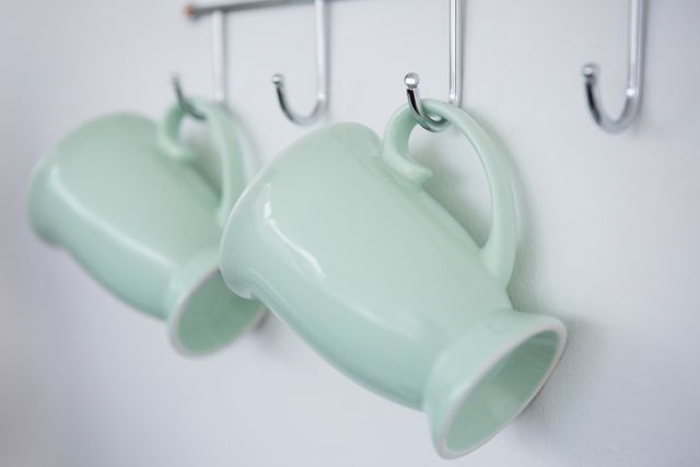 Close-up of two green ceramic mugs hanging on metal hooks against a white wall. Ideal for use in articles or blogs about kitchen organization, minimalist home decor, or ceramic kitchenware. Can also be used in advertisements for kitchen products or home improvement.