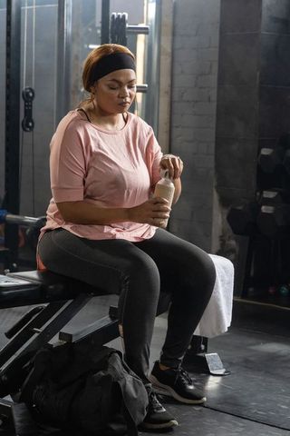 African american woman resting on gym bench hydrating from shaker between workouts