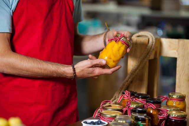 Market staff wearing red apron checking a pickle jar at a counter with various preserved jars. Ideal for use in articles about local markets, small businesses, organic food, and customer service in retail environments.