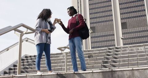 Young diverse professionals clinking coffee cups on urban steps near office tower