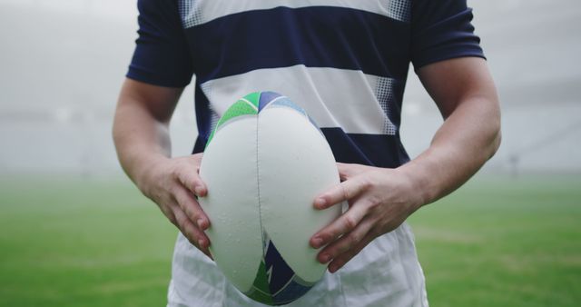 This image shows a rugby player holding a rugby ball on a grassy field, preparing for a game. It can be used to promote sports events, rugby tournaments, athletic wear, teamwork, physical fitness, and outdoor activities. The focused hands and ball capture the essence of preparation and readiness in sports.