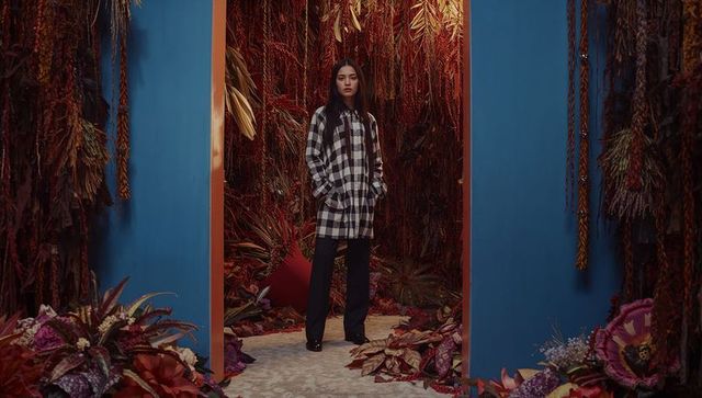 Model standing in blue portal with warm foliage wearing checkered coat and black trousers