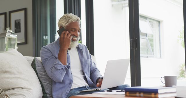 Senior man with grey beard working from home on his laptop while talking on the phone. Wearing casual clothing and sitting on the couch in a living room, he embodies a modern, relaxed remote work setting. Ideal for illustrating concepts like senior professionals, remote working trends, tech-savvy older adults, or work-life balance for seniors.